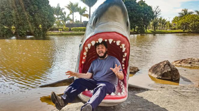 Beto Carrero World - O tubarão é um ótimo local para fotos divertidas Uma escultura de um tubarão de boca aberta sai das águas do lago ao fundo. O Coiso está sentado na escultura, dentro da boca do tubarão, com uma expressão aterrorizada