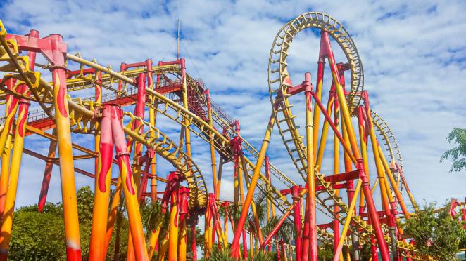 Brinquedos do Beto Carrero World Montanha russa radical FireWhip Trilhos de montanha-russa e pilares de sustentação nas cores laranja e vermelha cobrem todo o quadro em frente a um céu azul com muitas nuvens.