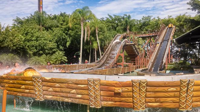 Brinquedos do Beto Carrero World Tchibum Surge à esquerda um barco cercado por respingos d'água com uma garota jovem sorrindo à frente. No restante da foto, o caminho traçado pelo barco durante o percurso da atração.