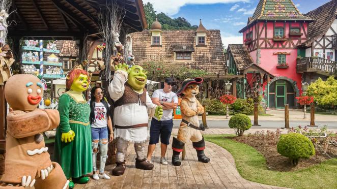 No parque Beto Carrero World voce pode encontrar os personagens de Shrek Biscoito, Fiona, Shrek e Gato de Botas posam e acenam ao lado de uma família em frente a edifícios com arquitetura clássica alemã