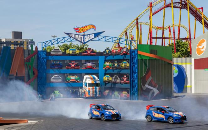 Beto Carrero World - Show Velozes e Furiosos Em primeiro plano, dois carros fazem manobras em frente ao cenário de uma garagem de Hot Wheels e um caminhão enquanto seus pneus soltam fumaça. Ao fundo, trilhos de montanha-russa erguem-se sobre o cenário.