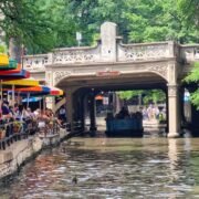 Mesas de restaurante com clientes sentados à beira do rio San Antonio, onde um barco com turistas passa sob uma ponte.