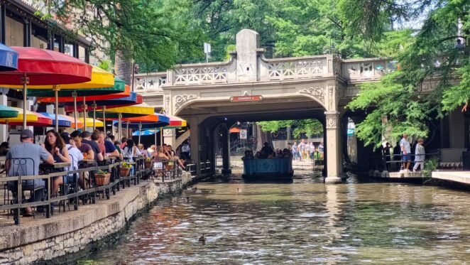 Mesas de restaurante com clientes sentados à beira do rio San Antonio, onde um barco com turistas passa sob uma ponte.